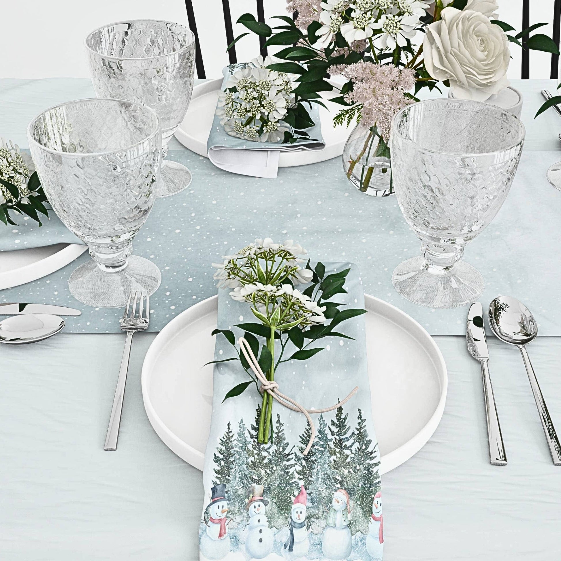  A dining table elegantly arranged with a white plate, silverware, and festive "Let it Snowmen" cloth napkin.